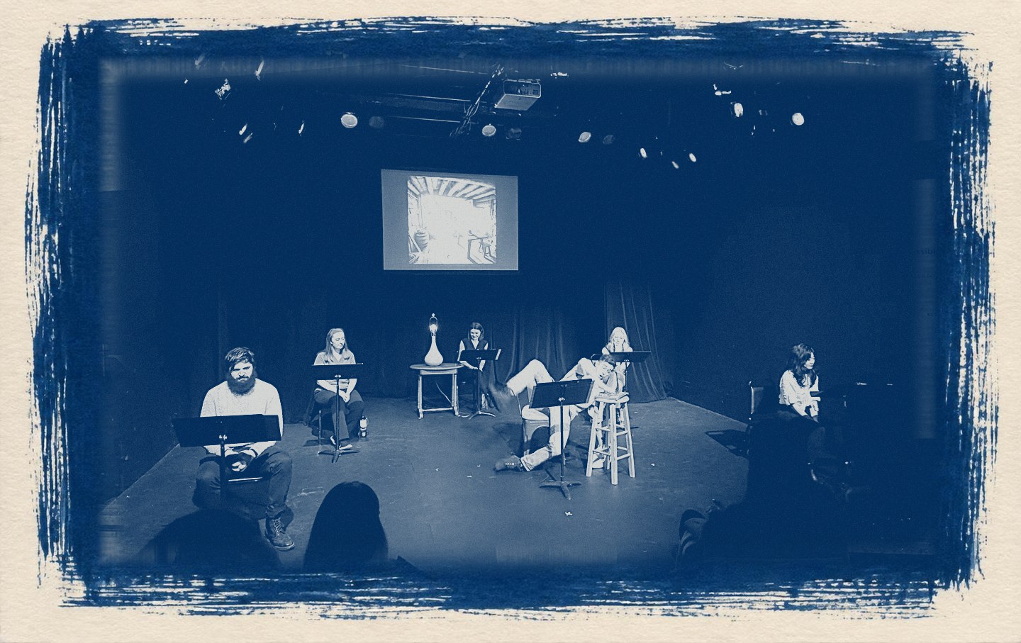 A cyanotype print of a staged reading in a black box theater: actors at music stands, one sprawled across a chair mid-scene, a projected cabin interior behind them and the audience in silhouette.