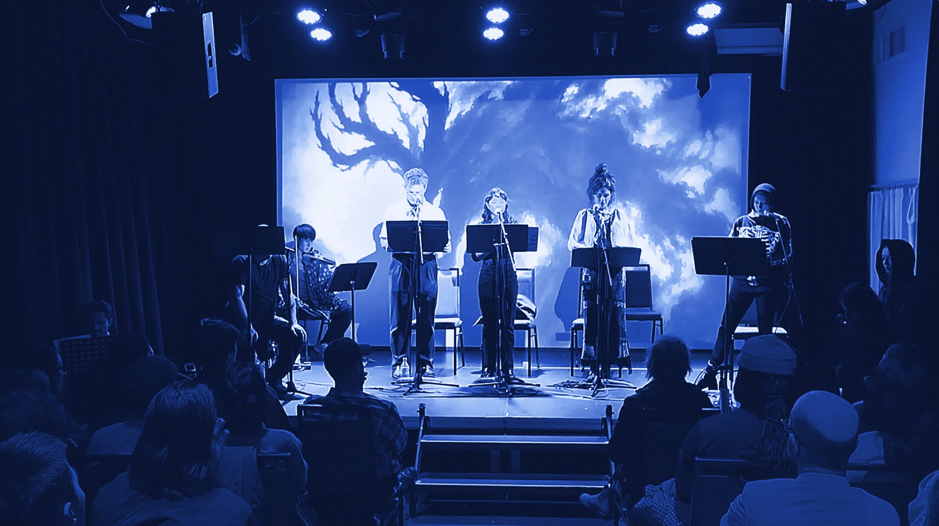 A staged reading: performers at music stands with musicians beside them, in front of a projected tree, as the audience watches.